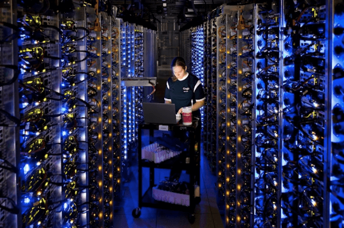 imagem em um dos data centers do Google com uma desenvolvedora fazendo manutenção em um computador que estava superaquecendo. Ela tem estatura mediana, está de camiseta preta e olhando para a tela de um notebook no centro da foto.