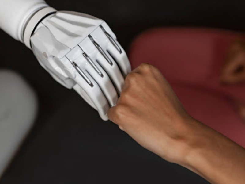 A imagem mostra uma cena simbólica e impactante: um cumprimento de punhos fechados ("fist bump") entre uma mão humana e uma mão robótica. Esse gesto transmite a ideia de parceria, colaboração e respeito mútuo entre humanos e inteligência artificial (IA).