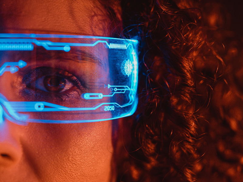 A imagem mostra o rosto parcial de uma pessoa, focando em um dos olhos, que está coberto por um óculos futurista com interface digital holográfica azul.