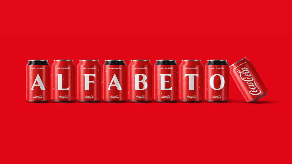 Oito latas de Coca-Cola vermelhas, alinhadas em um fundo vermelho intenso, formam a palavra 'ALFABETO' em letras brancas. O visual reforça a tipografia e o design icônico da marca, capitalizando na nostalgia e na familiaridade do símbolo.
