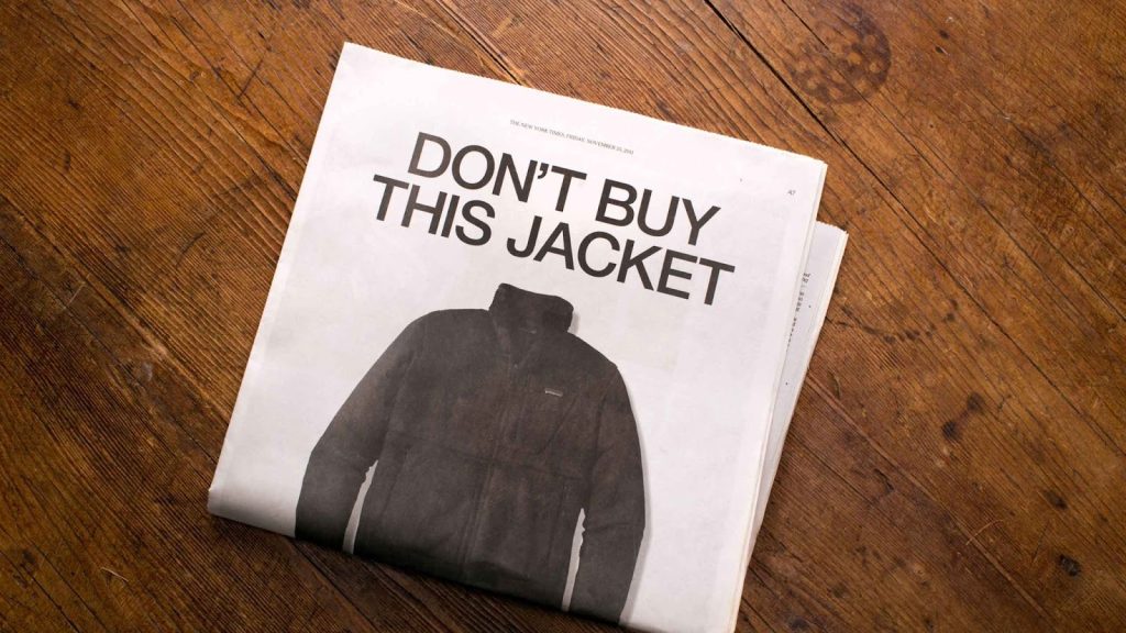 Foto de um anúncio de jornal da marca Patagonia. A manchete em letras grandes diz: 'DON'T BUY THIS JACKET' (Não Compre Esta Jaqueta). Abaixo, há a imagem de uma jaqueta de lã escura. O jornal está sobre uma superfície de madeira escura.