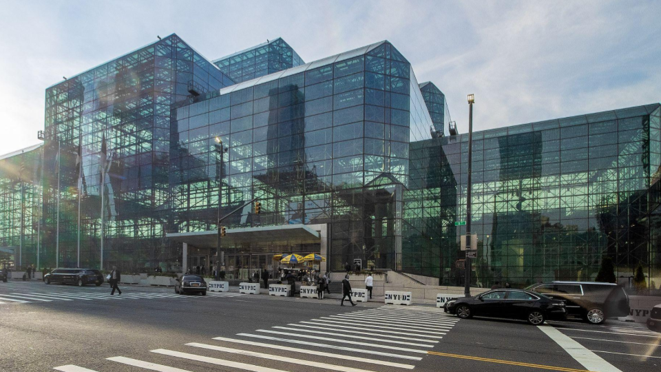 Fachada externa do Javits Center em Nova York. O edifício possui uma arquitetura moderna e imponente, composta inteiramente por painéis de vidro escuro e estruturas metálicas que refletem o céu e a luz do sol. Em frente ao centro de convenções, há uma rua movimentada com faixa de pedestres, alguns carros estacionados e pessoas caminhando na calçada sob a luz clara do dia.
