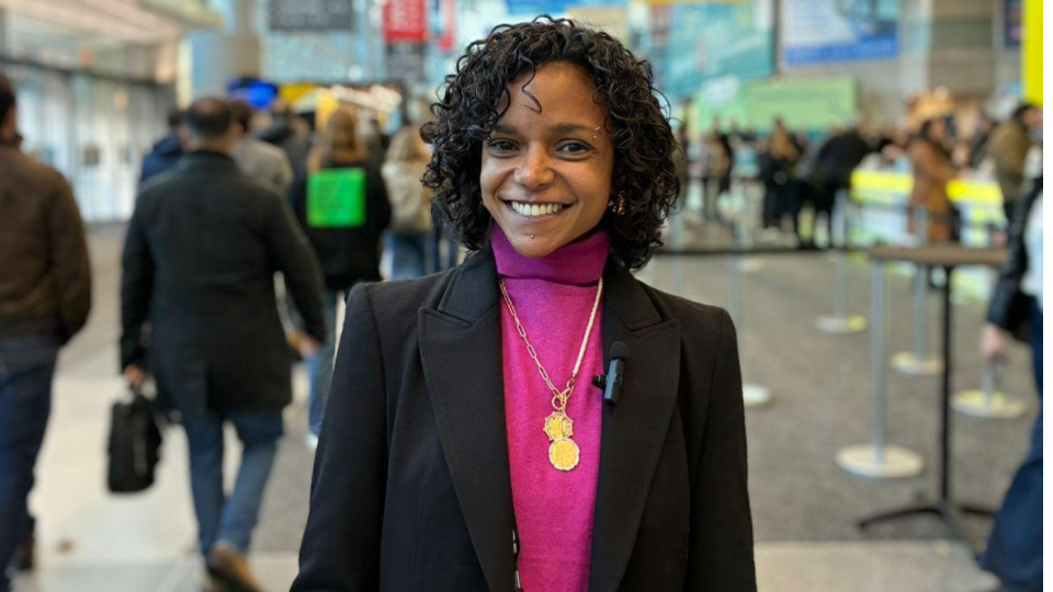 Carol Peres, CEO da Search One Digital, sorri para a foto no saguão movimentado da NRF Retail's Big Show. Ela é uma mulher de pele preta com cabelos cacheados curtos e escuros. Veste um blazer preto elegante sobre uma blusa de gola alta rosa vibrante, adornada com um colar de corrente dourada e pingentes. No blazer, nota-se um pequeno microfone de lapela acoplado. Ela carrega um crachá de identificação oficial do evento e segura um catálogo ou caderno claro. Ao fundo, o ambiente amplo do centro de convenções exibe sinalizações coloridas e diversas pessoas circulando.