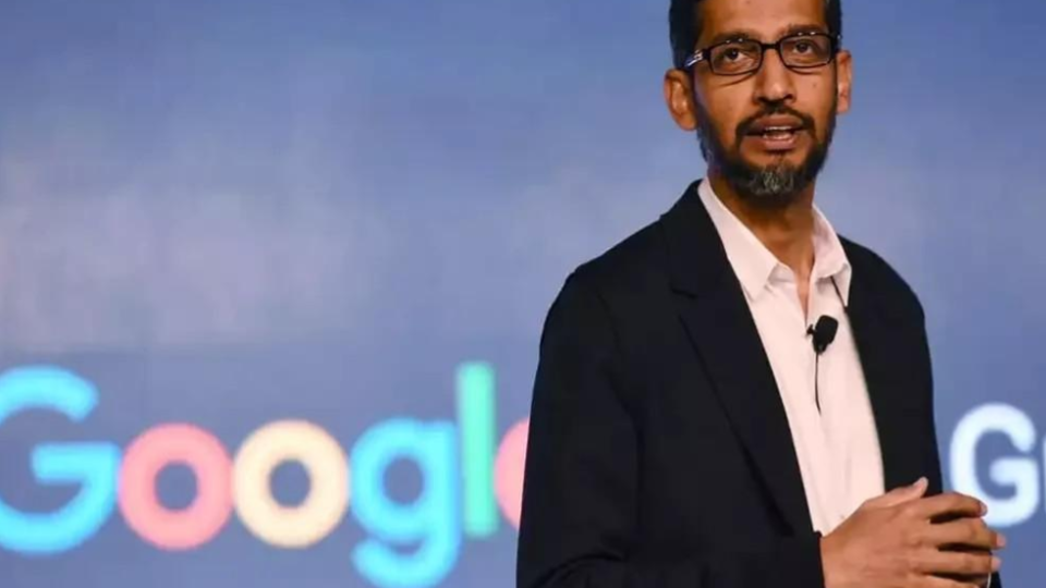 Fotografia de meio corpo de Sundar Pichai, CEO do Google, durante NFR 2026. Ele é um homem de pele retinta, usa óculos de armação escura, tem barba curta e grisalha e veste um paletó preto sobre uma camisa social branca levemente aberta no colarinho. Pichai está posicionado à direita da imagem, com as mãos gesticulando à frente do corpo. Ao fundo, há um painel azul levemente desfocado onde se vê parte do logotipo colorido do "Google".