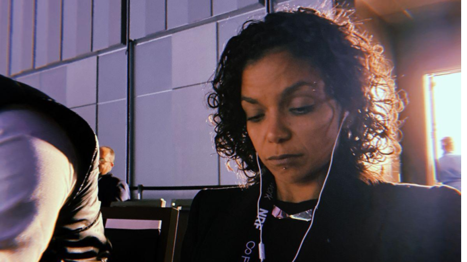 Carol Peres, CEO da Search One Digital, aparece concentrada em seu smartphone durante a NRF Retail's Big Show. Ela é uma mulher de pele preta e cabelos cacheados escuros, veste um casaco preto e utiliza fones de ouvido brancos com fio. No pescoço, carrega um crachá oficial do evento com os dizeres "NRF" e "Firework". A imagem possui um estilo cinematográfico com iluminação lateral e um carimbo de data retrô no canto inferior esquerdo.