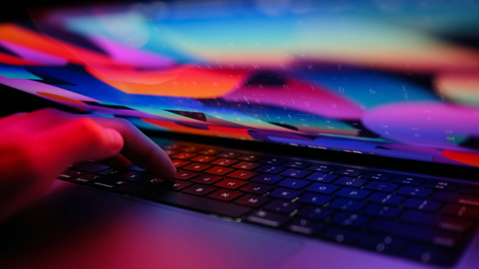 Vemos o teclado retroiluminado de um notebook escuro em ângulo diagonal. Uma mão humana aparece no canto esquerdo, com o dedo indicador pressionando uma das teclas.