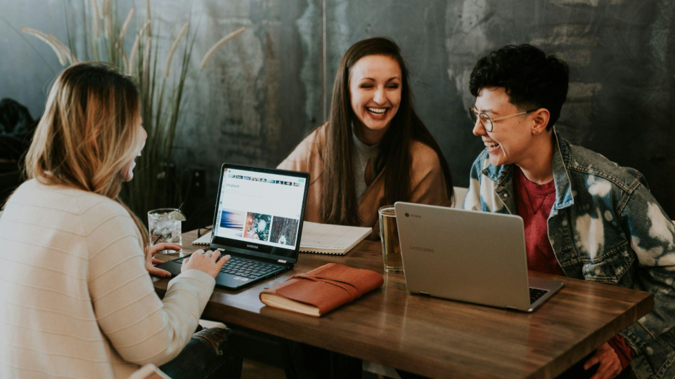 A foto retrata três pessoas reunidas em torno de uma mesa de madeira, conversando e sorrindo em um ambiente descontraído, como uma cafeteria ou espaço de coworking.