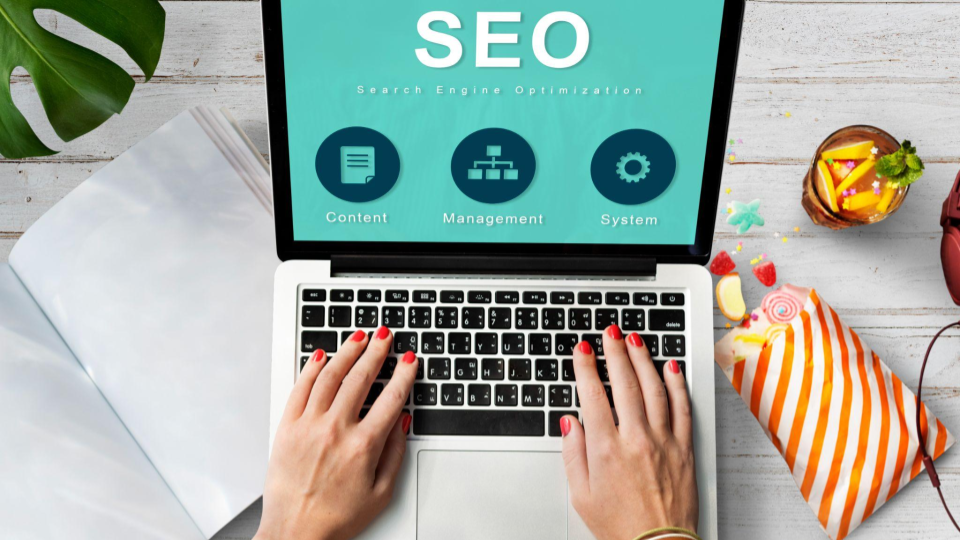 Um notebook prata aberto. Duas mãos femininas com unhas pintadas de vermelho estão posicionadas sobre o teclado. A tela exibe um fundo verde-menta com o texto destacado "SEO" (Search Engine Optimization) e três ícones circulares azuis com os rótulos Content (ícone de documento), Management (ícone de organograma) e System (ícone de engrenagem).