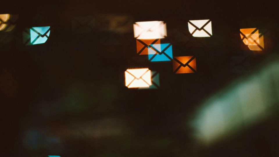 As luzes desfocadas formam o formato nítido de envelopes de e-mail/mensagens em tons brilhantes de branco, azul, laranja e amarelo, flutuando contra um fundo escuro e abstrato.
