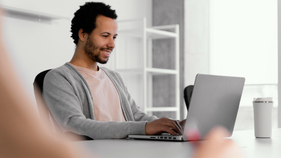 Um homem de barba, vestindo uma camiseta rosa-claro sob um casaco/cardigã cinza aberto. Ele está sorrindo enquanto olha para a tela do notebook.
