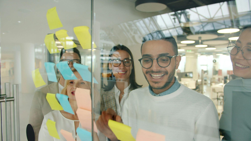 Um grupo de profissionais diversos trabalha de forma colaborativa em uma sessão de brainstorming, colando notas adesivas coloridas em um painel de vidro transparente.